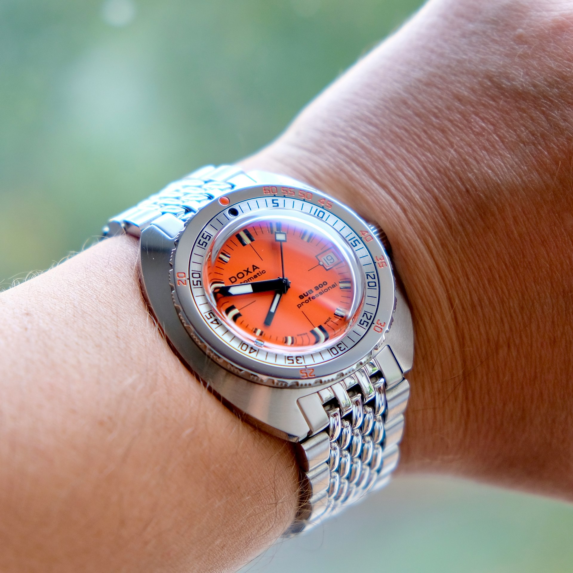 A silver wristwatch with an orange face and metal bracelet is worn on a person's wrist, displaying the time as 10:14.